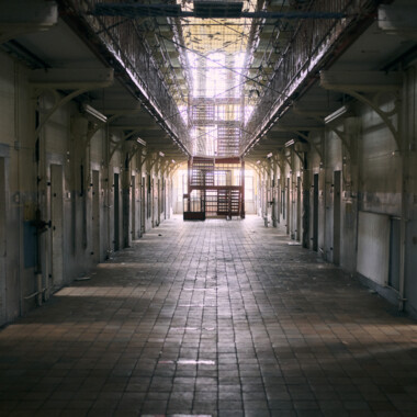 Eine Innenansicht der JVA Rennelberg. Ein langer, symmetrischer Gefängnisflur mit Zellentüren auf beiden Seiten. Der Boden ist gefliest. Am Ende des Gangs ist eine Treppe, dahinter Fenster. Durch das Gegenlicht ist das Foto relativ dunkel.